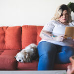Woman sitting on a red couch with her dog and reading a bog