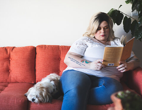 Woman sitting on a red couch and reading while a small white dog is sleeping next to her