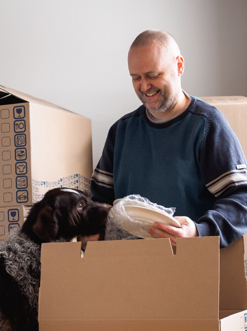 Man unpacking kitchenware from a moving box, a dog stands next to him