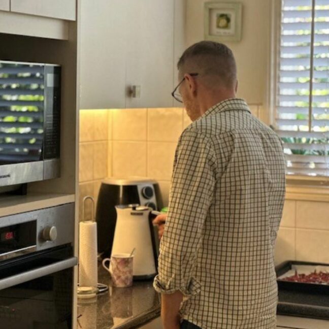 Man standing in a white kitchen using a kettle, with his back turned towards the camera