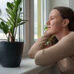 Woman with long blond hair looking thoughtfully out of the window. Her arms rest on the window sill.