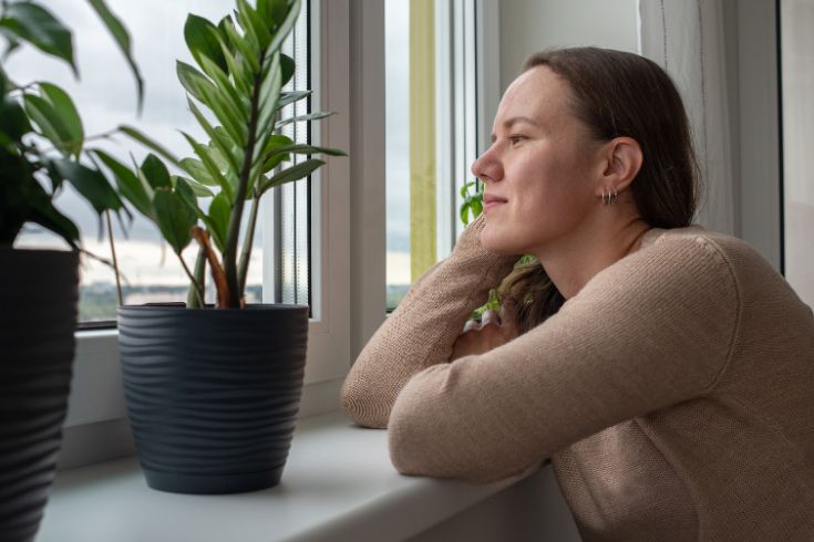 Woman with long blond hair looking thoughtfully out of the window. Her arms rest on the window sill.