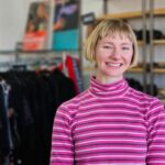 woman with short blond hair wearing a pink striped jumper standing in front of racks of clothing