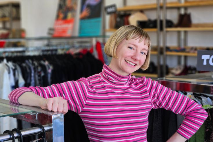 Jessie new store coordiantor at Sacred Heart Mission Op Shop Coburg Woman with short blonde hair and striped pink jumper leaning on rack of clothing