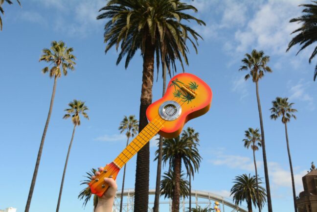 A ukelele is held up in the air with palm trees and the Luna park roller coaster in the background