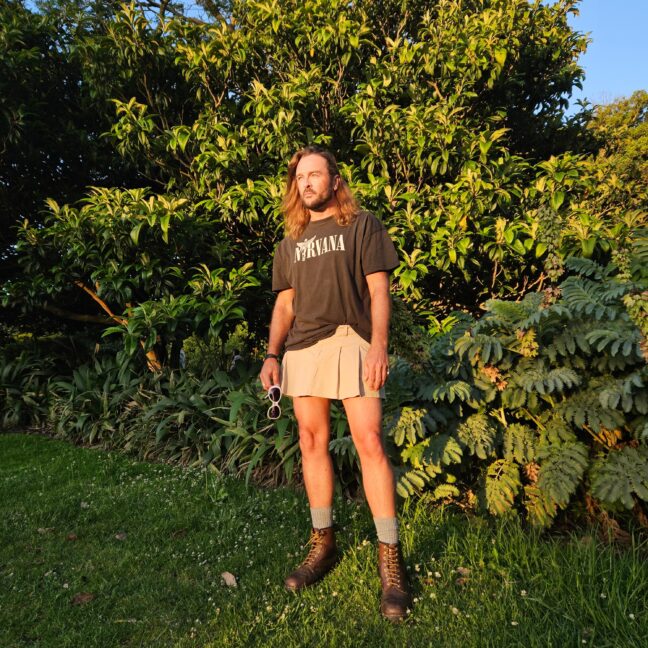 man wearing beige skirt, Nirvana t-shirt, combat boots standing in the afternoon sun surrounded by gardens.