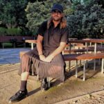 man sitting down in sun wearing skirt as part of a black outfit