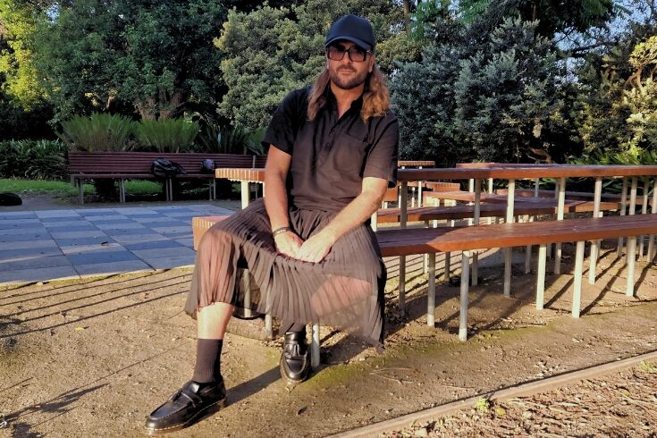 man sitting down in sun wearing skirt as part of a black outfit
